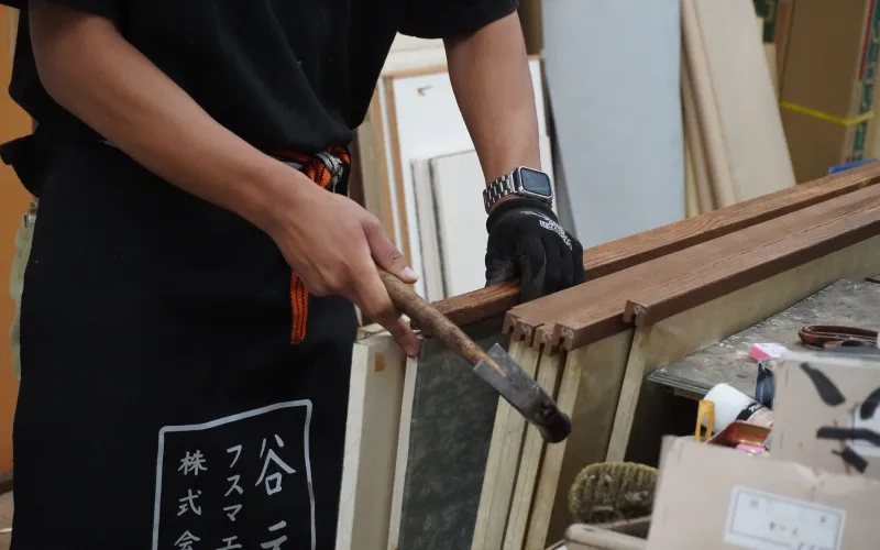 「谷元」と書かれたエプロンを着用した職人が、木槌を使って木材の調整作業をしている場面