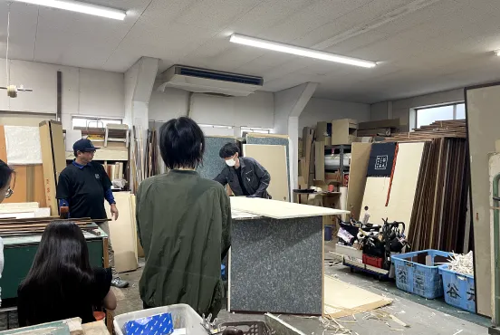 材料や道具が散らばった工房で、職人が板材を加工している光景