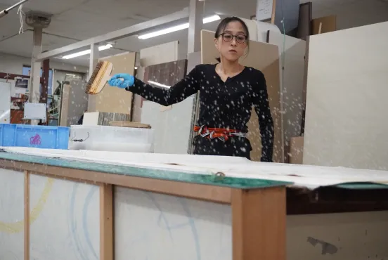 女性職人が作業台で水を飛ばしながら布地を伸ばす作業をしている動きのある場面