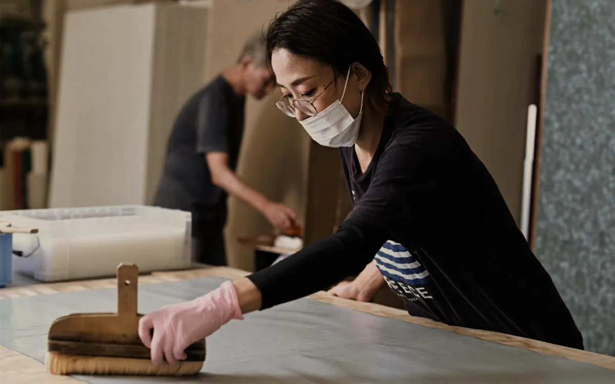 「室内ドア通販_和室リフォーム本舗」の敷地内にある工場風景。女性職人が刷毛でノリを塗っている風景